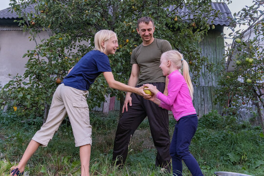 Happy father and two blond children, a boy and a girl, engaging in a playful tug of war over a fresh apple, enjoying family time in a rustic backyard setting