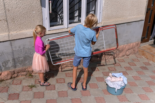 Young boy and girl working together, preparing a clothes drying rack on a paved patio outside their home, with a basin of laundry waiting to be hung under the bright sun