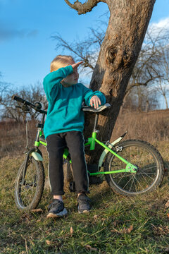 Young girl beside a green bicycle and old tree, shielding his eyes as he gazes into the distance across a sunlit rural field, dreaming, exploring and planning adventure