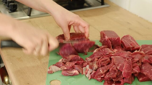 Close up of female hands cutting raw beef meat with a sharp knife on a cutting board in the kitchen, preparing fresh ingredients to cook a delicious homemade dinner. Red steak fillet dicing process