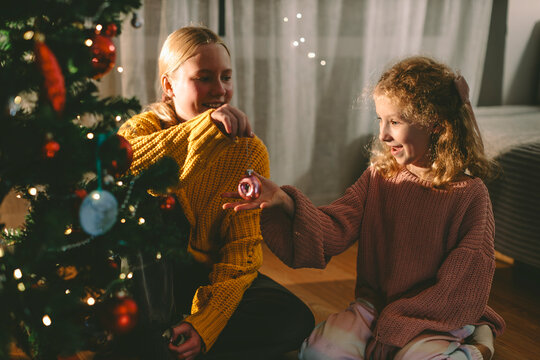 Sisters decorate a Christmas tree indoors with a pink bauble
