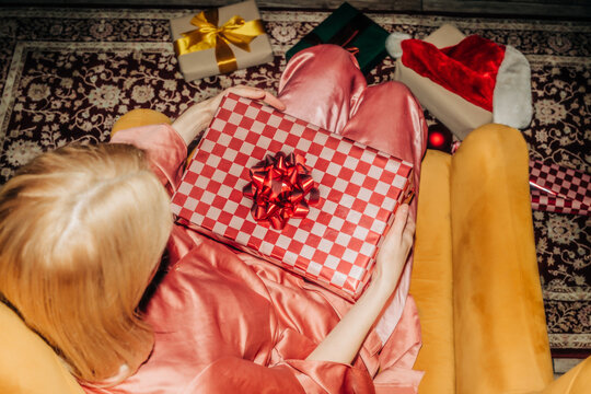 Woman in sleepwear opens a Christmas gift in a yellow armchair at home