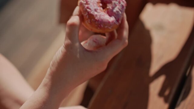 Young child receives and examines pink frosted donut with sprinkles showing curiosity about treat. scene is set outdoors on wooden surface under natural light focusing closely on interaction