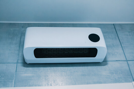 White ceramic electric space heater on tiled floor against a bathroom wall. Home heating appliance for product or retail use.
