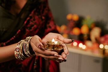 Obraz premium Cropped hands of Indian young adult woman holding lit brass oil lamp during traditional festival celebration. Visual content for seasonal marketing campaigns and cultural greeting designs