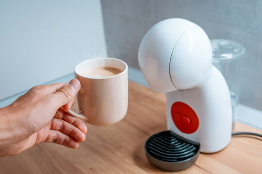 Hand holding a fresh latte mug next to a compact pod coffee machine on a wooden kitchen counter. Home brewing concept.