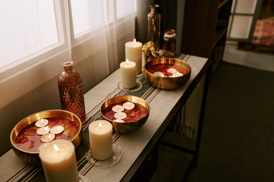 High angle shot of lit candles and floating tealights in brass bowls with flower petals on table indoors. Still life composition suitable for wellness, spa, and festival marketing