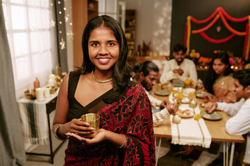 Obraz premium Portrait of smiling Indian young adult woman holding cup during festive dinner with family at home. Image suitable for holiday marketing campaigns and cultural celebration promotions