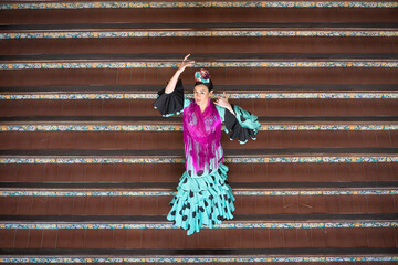 An Andalusian woman wearing a turquoise flamenco dress with black polka dots and a purple shawl, with flowers in her hair, dancing flamenco on a staircase in Seville’s Spain Square. Elevated view. © Manuel