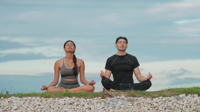 Two people meditate in lotus pose. They sit on grass beside white stones. Sky is soft with scattered clouds. They appear calm and focused