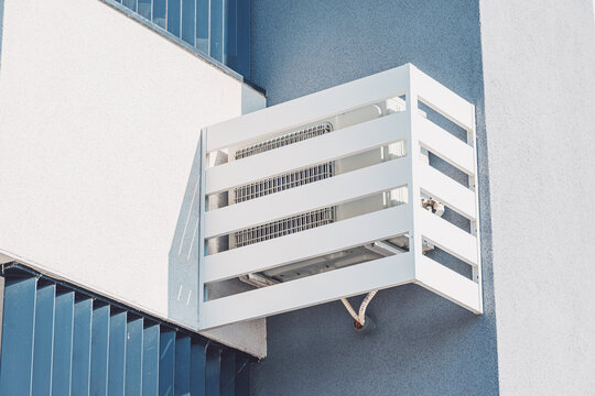 Slatted white cage enclosure protecting an outdoor air conditioning unit mounted on a building exterior wall. Property maintenance concept.