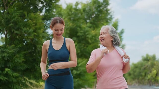 Two women jog side by side through a green park. Younger woman in blue athletic gear walks beside an older woman. The older woman wears pink