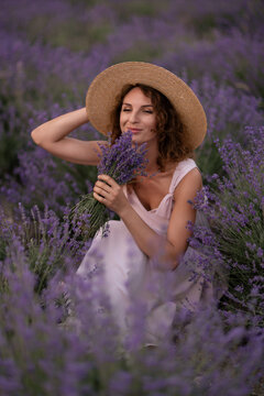Lavender bouquet held by young woman wearing straw hat crouching in purple Lavandula field at sunset