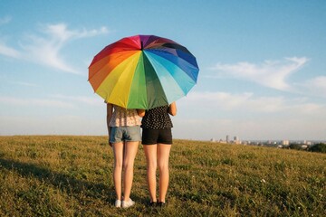 Obraz premium Two young women stand in grass field under large rainbow umbrella. Back view of girls holding pride flag colored parasol. Look at city skyline on horizon during sunset. LGBTQ community concept of