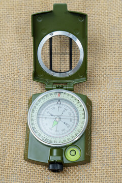 Green military style lensatic compass open on a textured burlap background, displaying navigation and orientation tools
