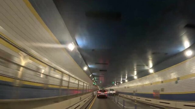 Driving inside a traffic tunnel with cars, lane lines, and ceiling lighting in dashcam view