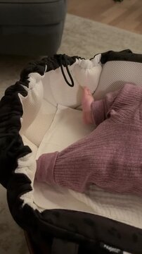 Baby's bare feet and legs in a purple ribbed romper kicking playfully inside a cozy bassinet at home, evoking early childhood, tenderness and new parent life