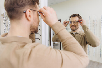 Obraz premium Man trying new frames in an optical shop, checking his look in the mirror for improved vision and style