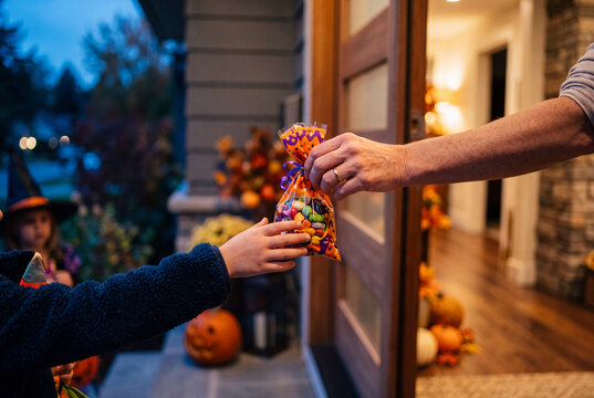 Primo piano di una mano adulta che consegna un sacchetto di caramelle colorate a un bambino in costume sulla soglia di casa, al crepuscolo, con zucche intagliate e decorazioni autunnali sfocate.