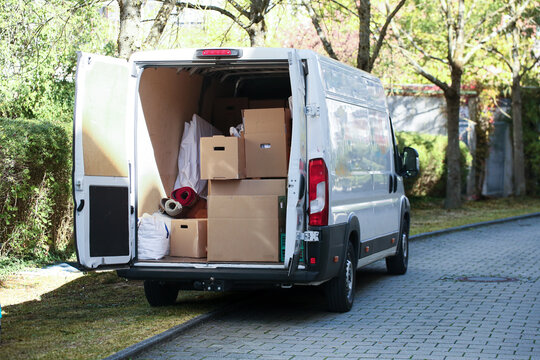 Wei&szlig;er Transporter mit ge&ouml;ffneten Heckt&uuml;ren, beladen mit Umzugskartons und M&ouml;beln. 3/4-Ansicht, geparkte Stra&szlig;e, B&auml;ume, Umzug, Logistik.