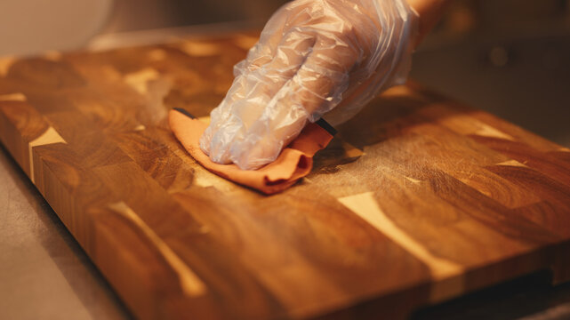 Close-up of a gloved hand rubbing hard wax oil onto an acacia wooden cutting board with a cloth for wood protection and care. This process restores the natural grain texture of the wood surface.
