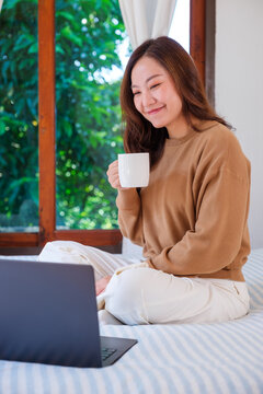 Vertical portrait image of a woman working on laptop computer and drinking coffee while sitting on a bed at home