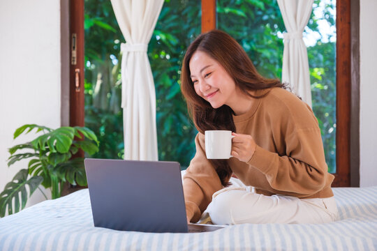 Portrait image of a woman working on laptop computer and drinking coffee while sitting on a bed at home