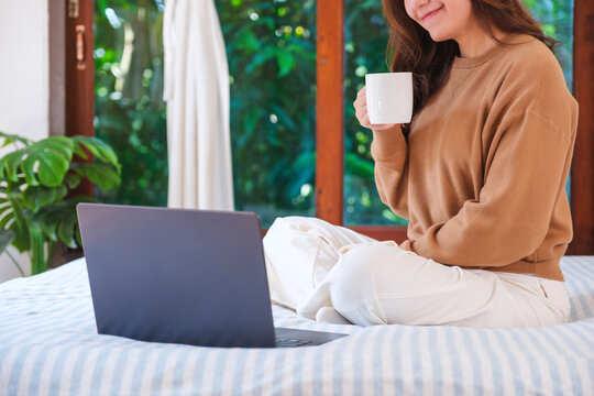 Closeup image of a woman working on laptop computer and drinking coffee while sitting on a bed at home