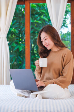 Vertical portrait image of a woman working on laptop computer and drinking coffee while sitting on a bed at home