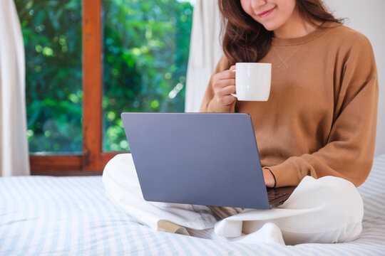 Closeup image of a woman working on laptop computer and drinking coffee while sitting on a bed at home