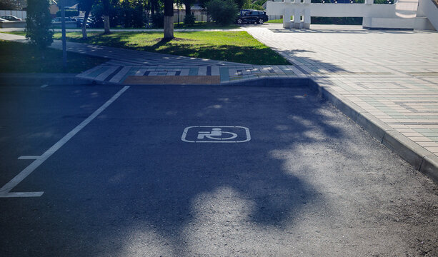 Accessible parking space marked with white wheelchair symbol painted on asphalt road next to sidewalk ramp leading to public square on sunny day