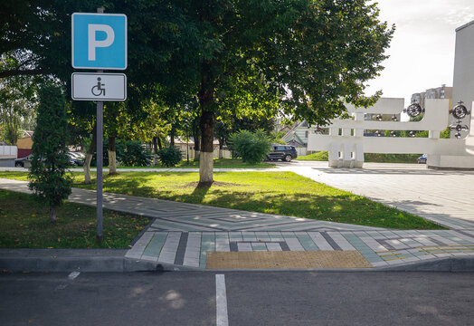 Blue accessible parking road sign standing next to sidewalk ramp with tactile paving blocks near public plaza on sunny day