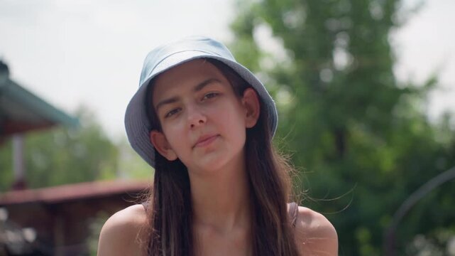 Young woman with long hair sits outdoors wearing light blue bucket hat and sleeveless top. background features greenery trees part of building structure like awning or porch roof
