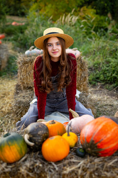 Young Woman with Pumpkins at Autumn Farm. Young woman in a straw hat sitting among colorful pumpkins and hay in a rustic autumn setting.