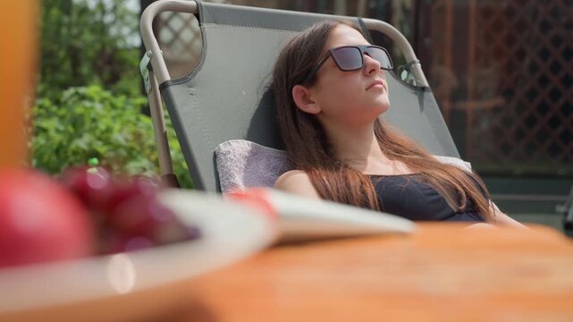 Young woman relaxes in sunlit outdoor setting wearing sunglasses and reclining on chair. plate of fruit is visible nearby with greenery softly blurred in background to emphasize her calm