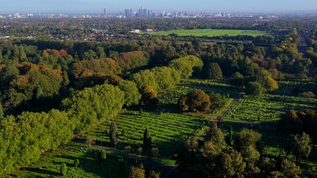 Sunlit aerial video looking over tree-lined avenues and headstone sections of Manchester Southern Cemetery toward the Manchester city centre skyline.