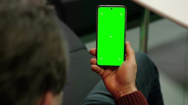 Man holding smartphone vertically with green screen chroma key and tracking marks indoor. Over the shoulder closeup view of male user looking at cell phone isolated blank template. Landscape footage.
