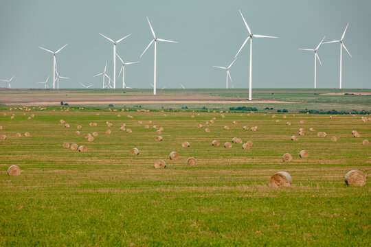 Wide rural landscape with rolled hay bales and modern wind turbines under a clear sky, blending agriculture and renewable energy.