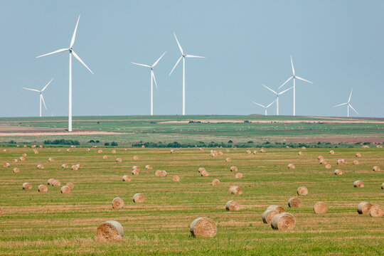 Modern wind turbines towering over a rural landscape with hay bales scattered across green farmland under a clear blue sky.