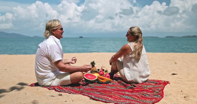 Mature couple enjoy time together relaxing on a sandy beach with a red picnic blanket and fresh tropical fruit, man and woman looking at each other, serene vacation day in Ko Samui Island, Thailand.