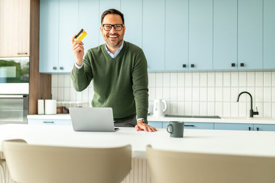 Man smiling and showing a credit card while standing in a modern kitchen, using a laptop for online shopping and secure payment transactions