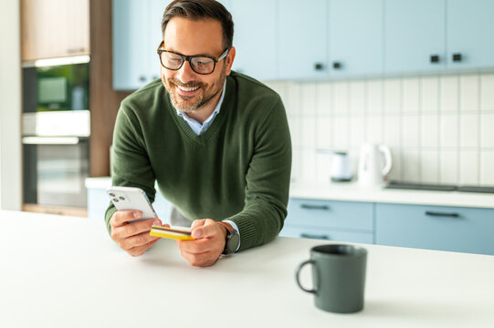 Man making an online purchase using a credit card and smartphone in a modern kitchen, confidently managing personal finances and e commerce transactions from home