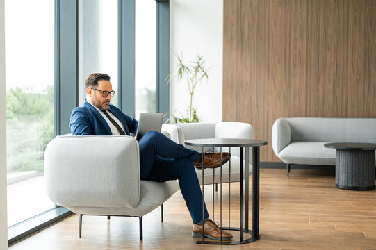 Mature businessman in a suit reviews financial and legal documents on a laptop while seated in a modern office lounge, focused and planning company strategy