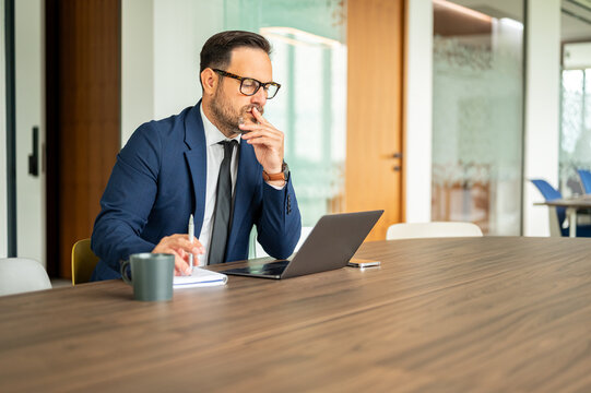 Businessman concentrating while working at a conference table, analyzing data on a laptop and taking notes during an important business meeting in a corporate office