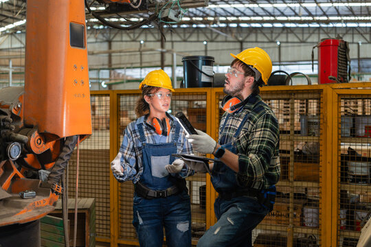 Gesturing, a woman in the carpentry workshop watches her colleague hold a tablet and shine a flashlight on a robot.