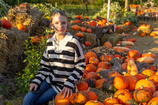 Young woman sitting among colorful pumpkins and hay bales in an autumn garden, warm sunlight creates a vibrant seasonal harvest scene.