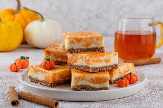 Pumpkin marble cheesecake bars on a white plate with mini pumpkin decor and tea, styled in autumn tones. Cozy fall dessert perfect for Thanksgiving or Halloween