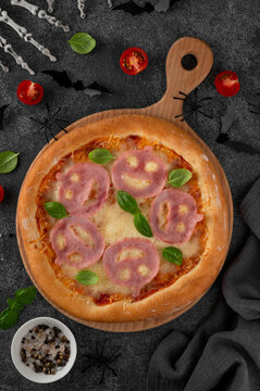 Halloween pizza with smiling ham faces, basil leaves and spooky decorations on a dark background, creative seasonal food concept for parties, menus, and social media. Top view. Copy space.
