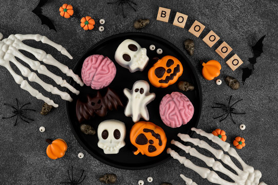 Halloween themed dessert. Mini cakes in colorful chocolate glaze in the shape of a ghost, brain, skull, and bat on a black plate on a concrete background. Top view, copy space.
