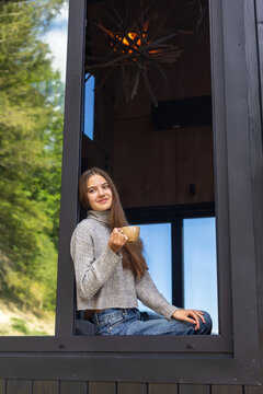 Young Woman Drinking Coffee by Cabin Window. Young woman holding a cup of coffee while sitting by a large window in a modern cabin surrounded by nature.